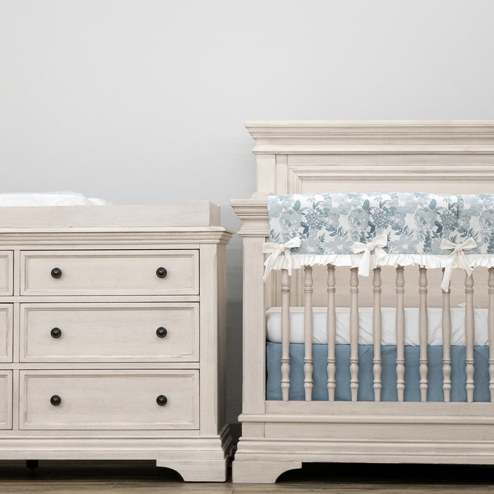 White nursery furniture set with crib and dresser against a light gray wall.