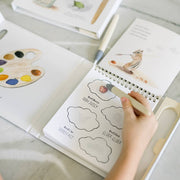Open book with coloring pages and a hand holding a pen on a light surface.