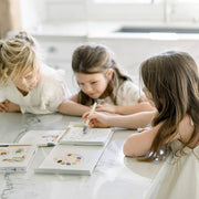 Three children sitting at a table with books and markers in a bright room.